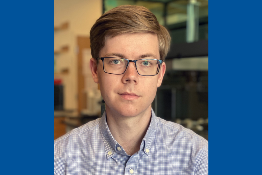 Pictured is Dr. Connor Coley, PhD, a man smiling at the camera with short, light brown hair and glasses, wearing a light blue checkered button-up shirt. The background is a modern laboratory.